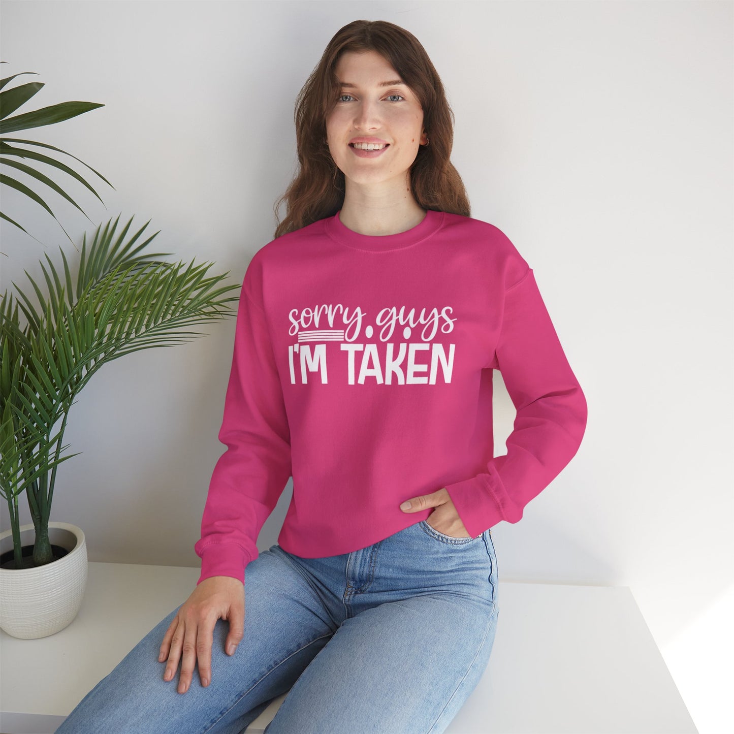 Woman wearing a pink sweatshirt with 'Sorry guys I'm taken' text, sitting on a white surface with a plant in the background.