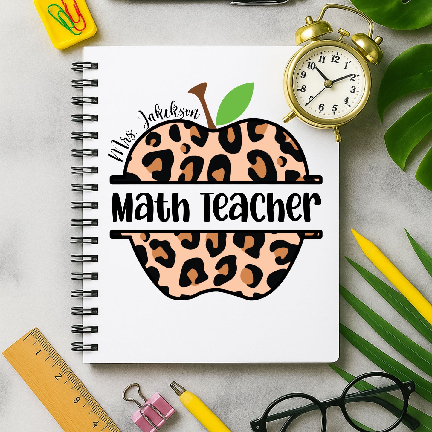 Notebook with leopard print apple design labeled 'Math Teacher' on a desk with stationery items.