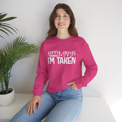 Woman wearing a pink sweatshirt with 'Sorry guys I'm taken' text, sitting on a white surface with a plant in the background.